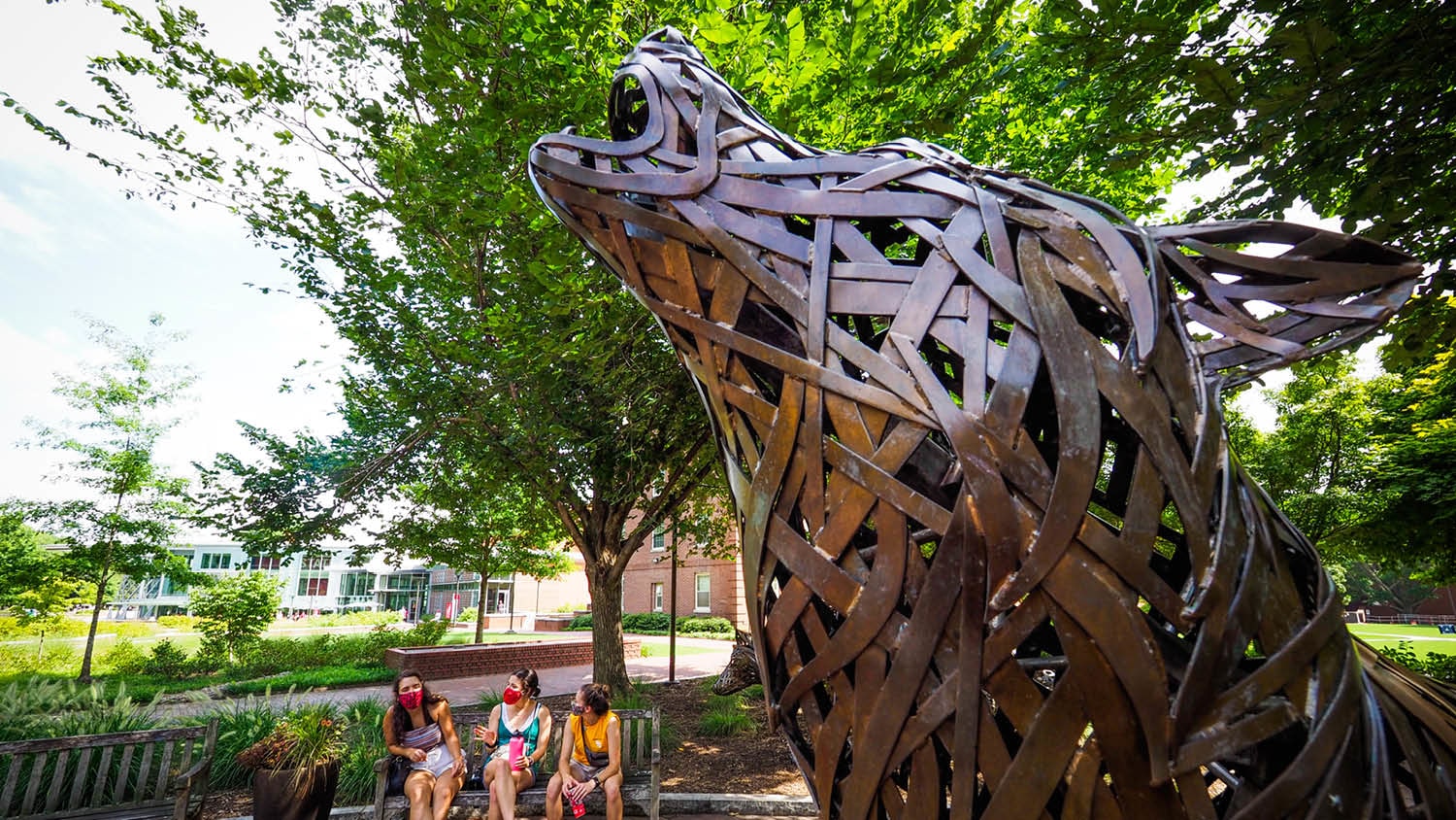 copper wolf howling in wolf plaza with students in the background sitting on a bench